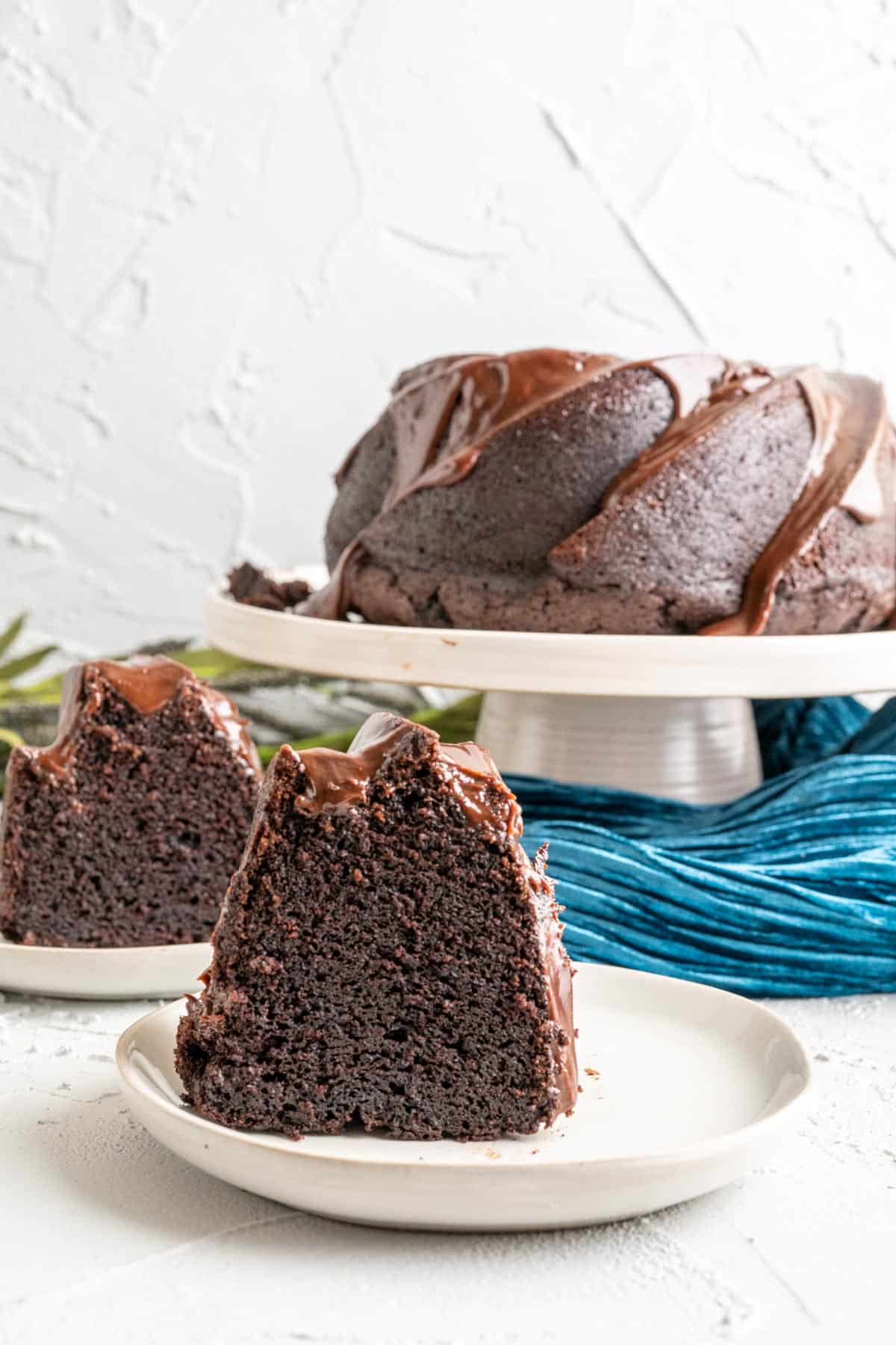 two pieces of dark chocolate bundt cake on white plates with the cake in the backdrop.