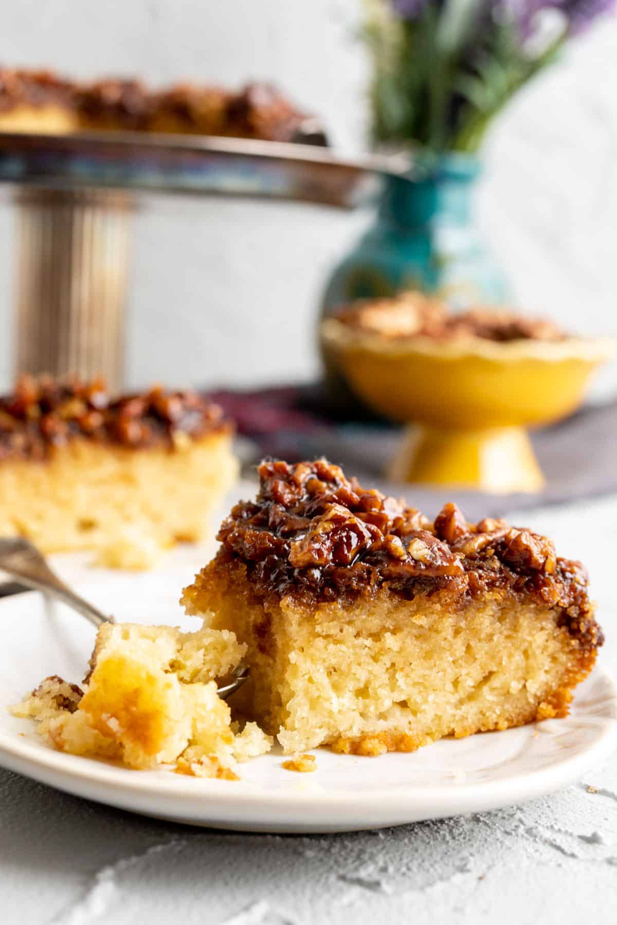 Slice of pecan upside down cake in a small white plate with a cake stand in the backdrop.