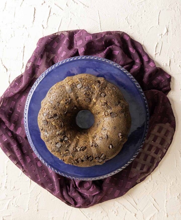 overhead shot of banana chocolate chip bundt cake sitting in a cake stand over a peach table with purple cloth.