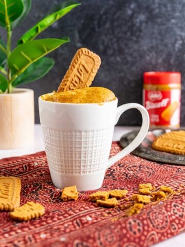 biscoff cake in a white mug with biscoff cookies spread around.