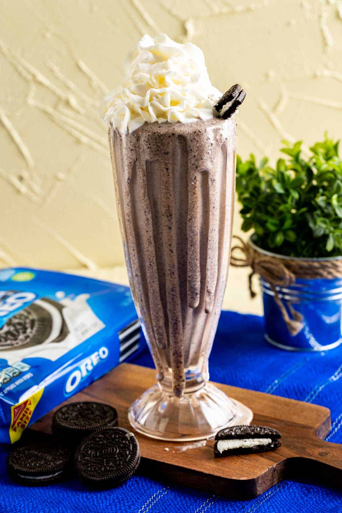 tall glass of oreo banana milkshake placed on a small wooden plank on a blue table cloth.