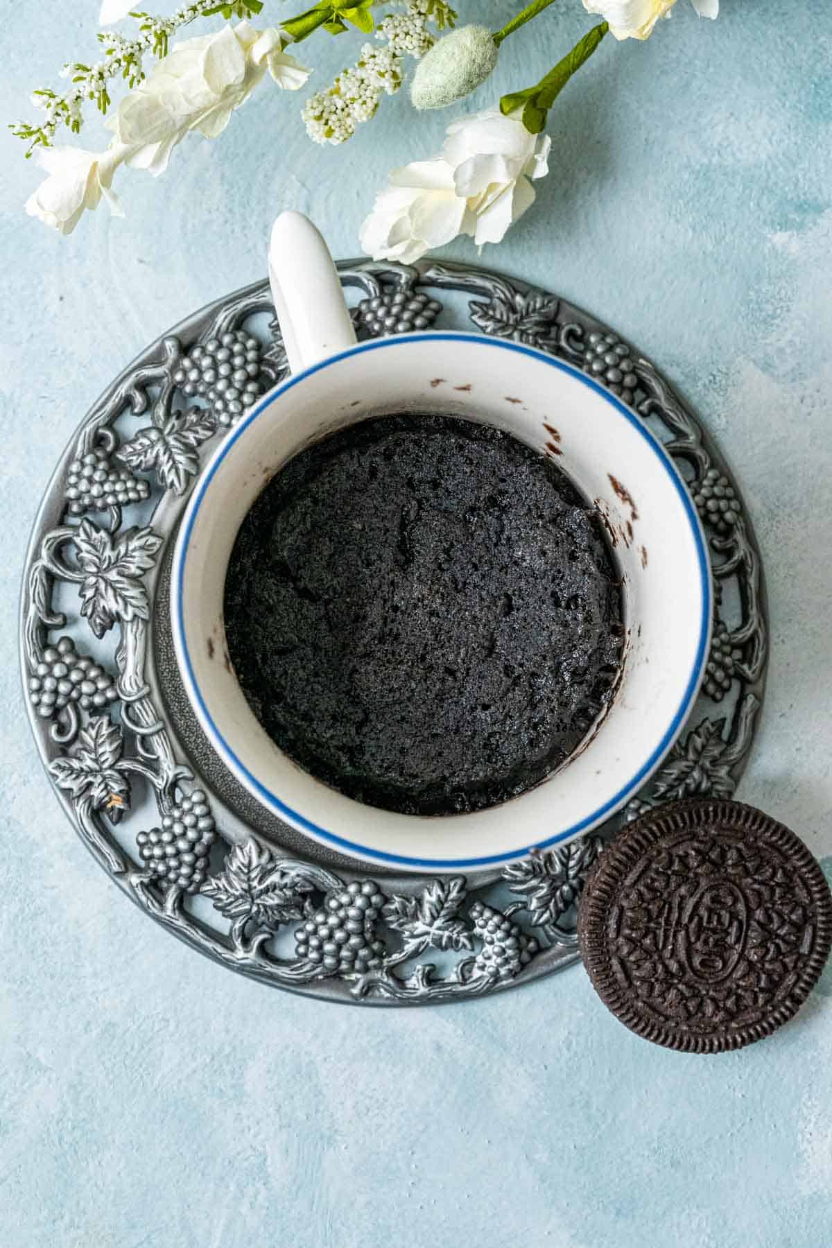Oreo mug cake in a white cup placed on a blue table with an oreo cookie on the plate.